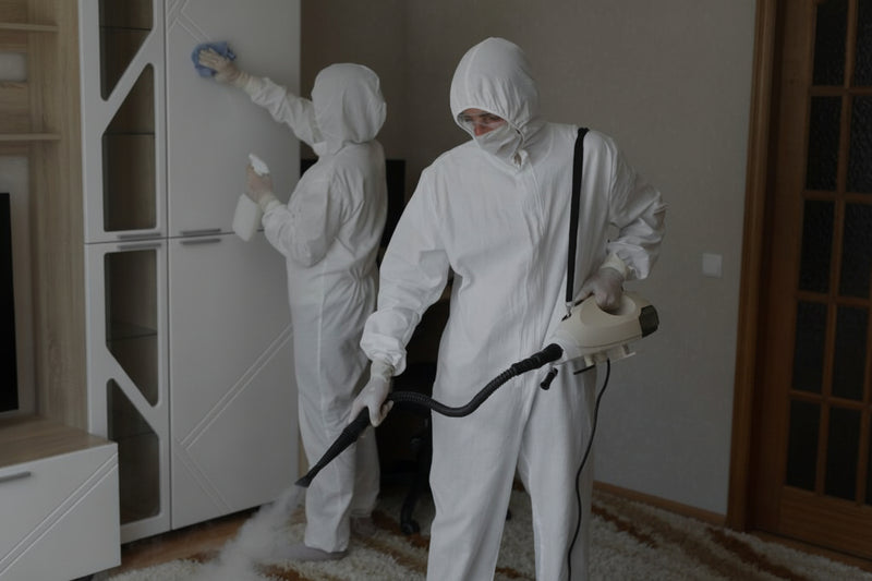 Two people in white protective suits cleaning a room with a vacuum cleaner.