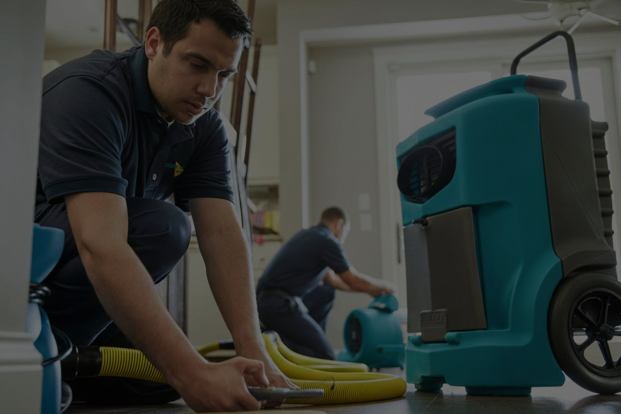 Person using a teal and black pressure washer in a room with another person in the background.