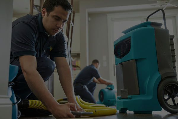 Person using a teal and black pressure washer in a room with another person in the background.