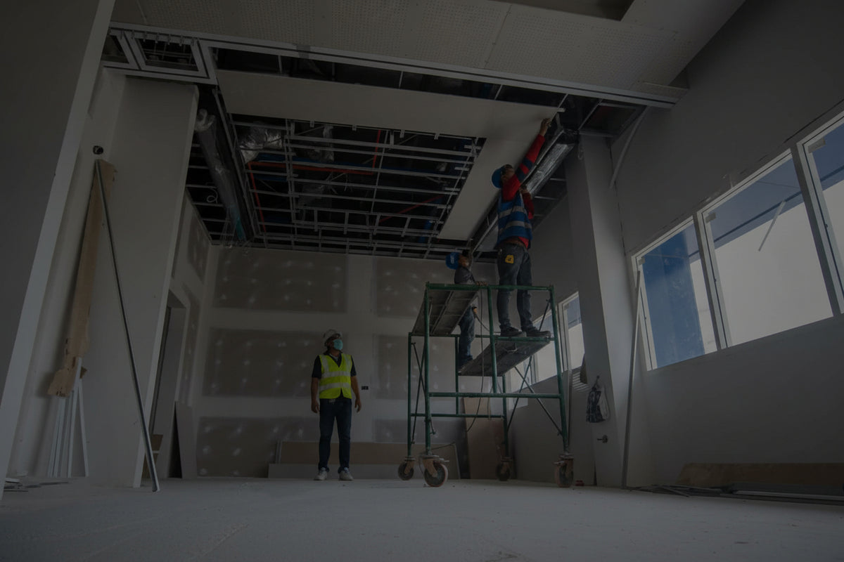 Two workers installing ceiling panels in a large room with large windows.