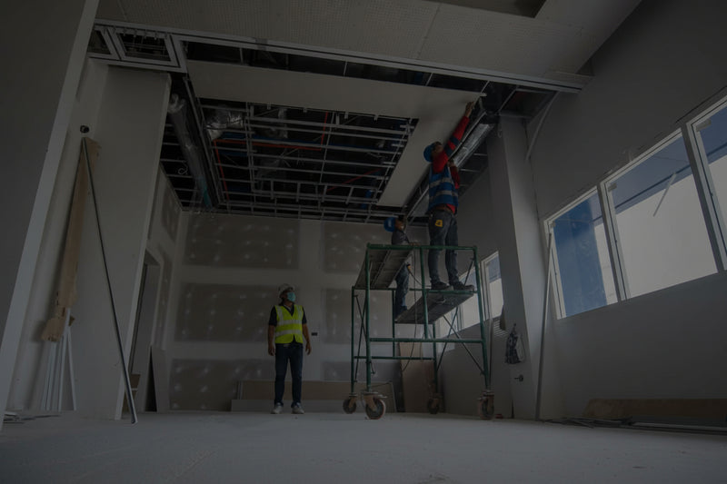 Two workers installing ceiling panels in a large room with large windows.